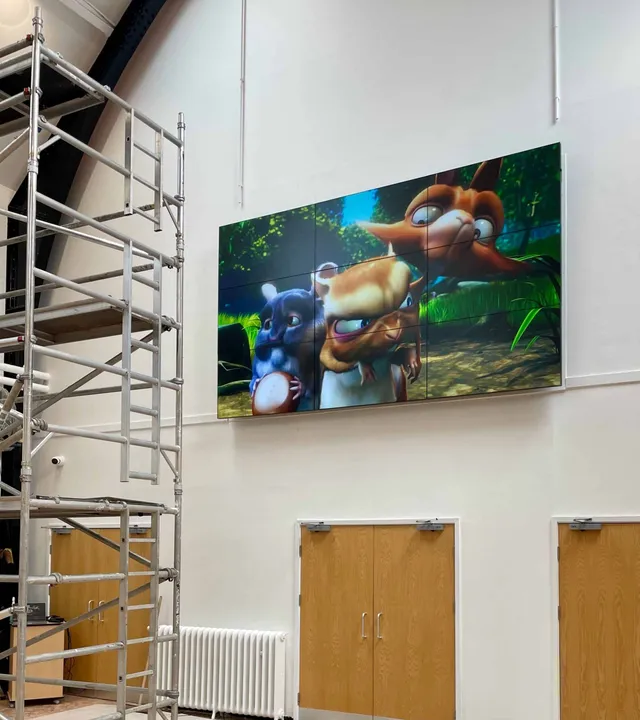 Large display showing a video game character, installed high on a venue wall beside access scaffolding